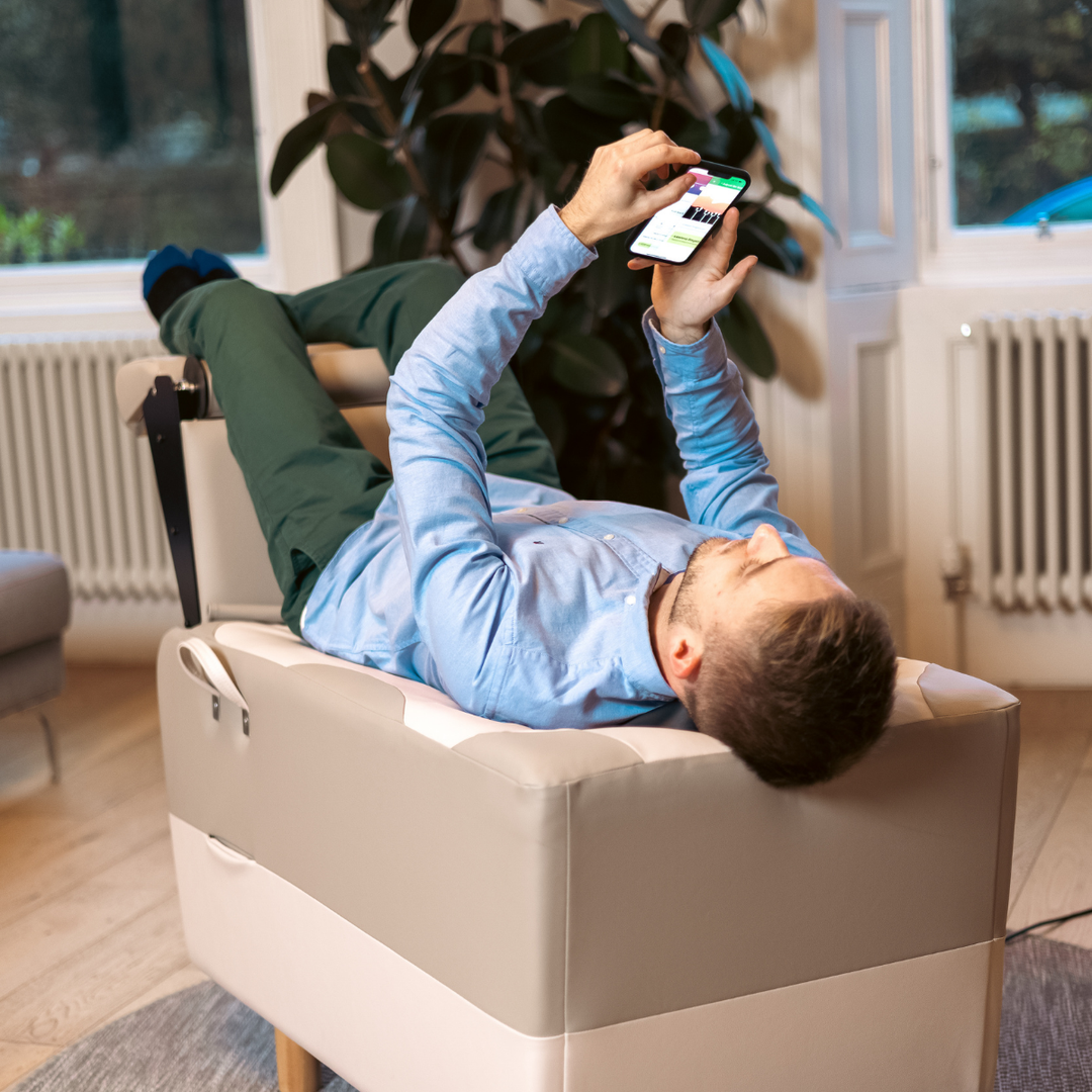 Man lying fully clothed on a BackHug robotic back therapy bed with his legs raised, scrolling his phone